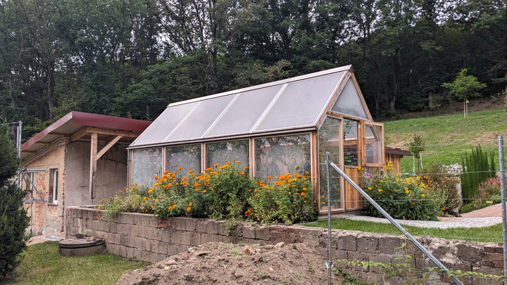 Ein langes Lucama-Gewächshaus auf einem gemauerten Steinsockel, davor ein Beet mit Blumen