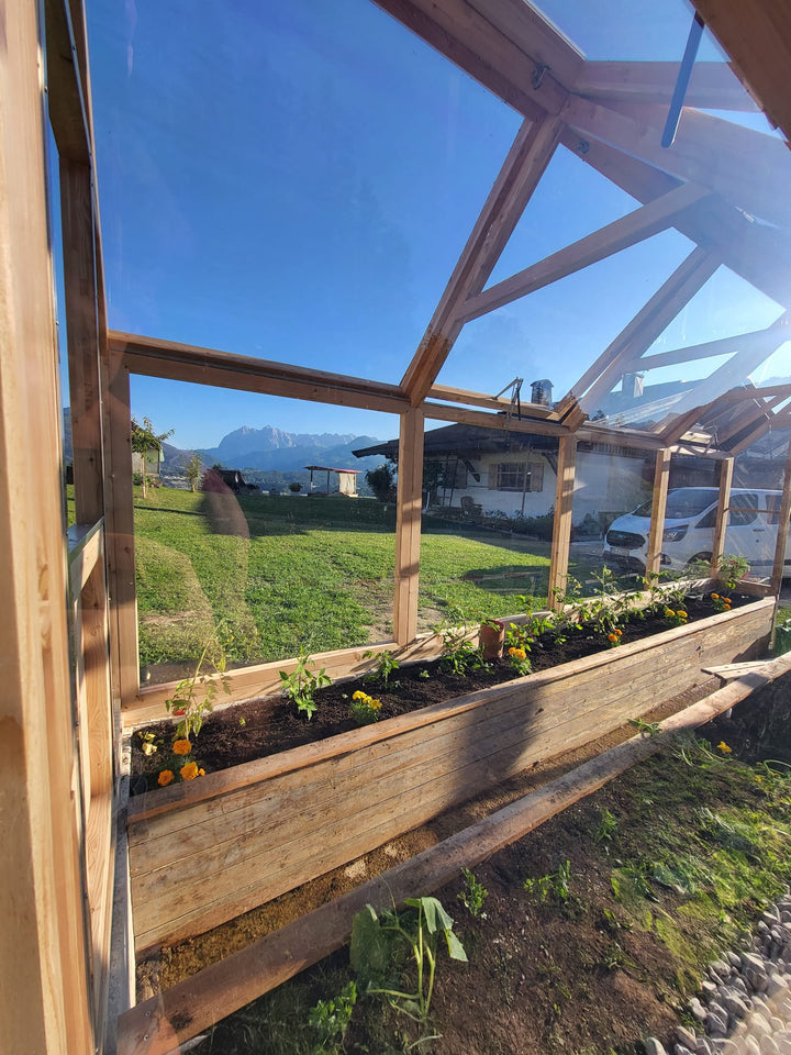 Blick aus einem Lucama-Gewächshaus über ein bepflanztes Hochbeet hinaus auf Wiesen und Berge